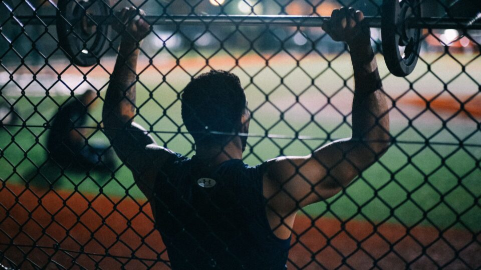 Man lifting a barbell outdoors at night behind a chain fence, emphasizing strength and fitness.